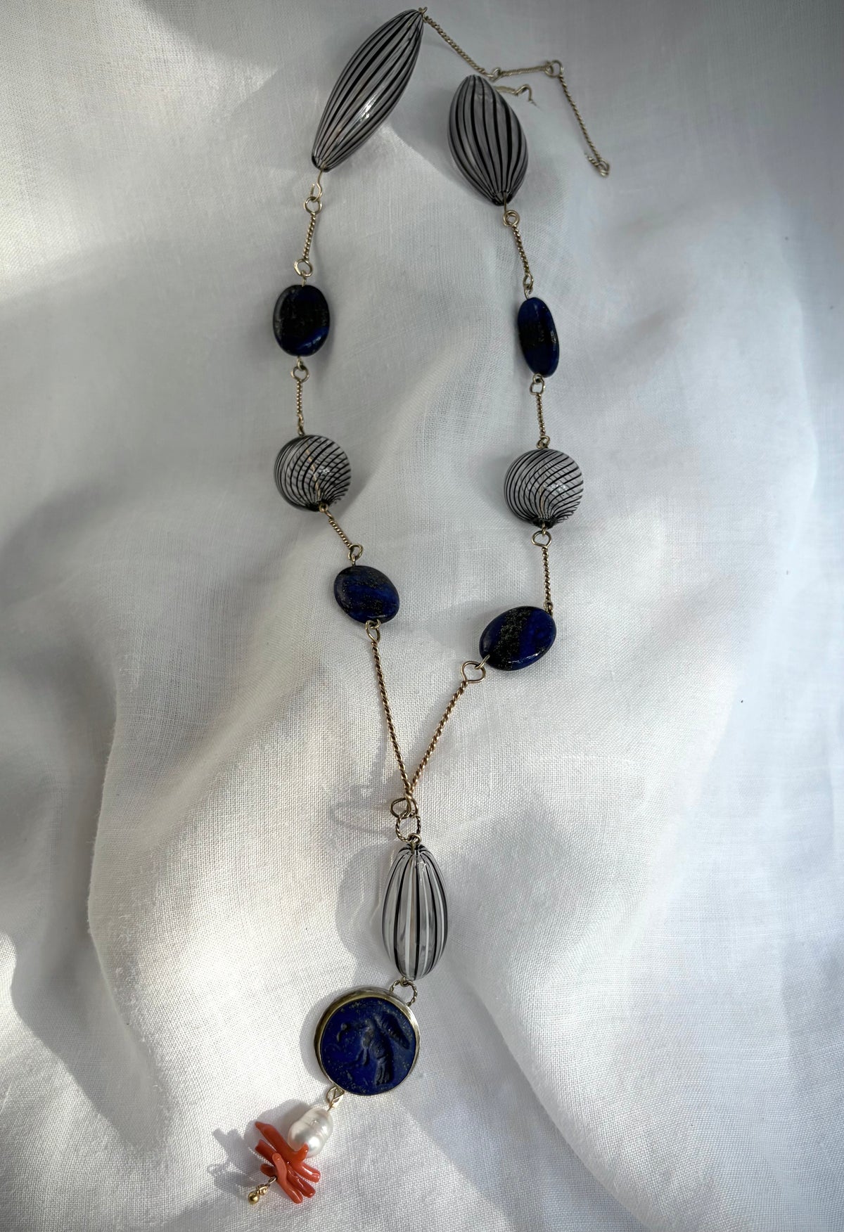A necklace twists diagonally across a white fabric-covered surface. The twisted gold wire is punctuated by large, oval, clear glass beads with black stripes and round ones with swirling parallel lines. In between are gold-flecked lapiz lazuli beads, with a focal pendant that features a lapis carved with a bird with a snake in its beak. A South Sea baroque pearl, natural coral, and tiny gold beads complete the necklace.