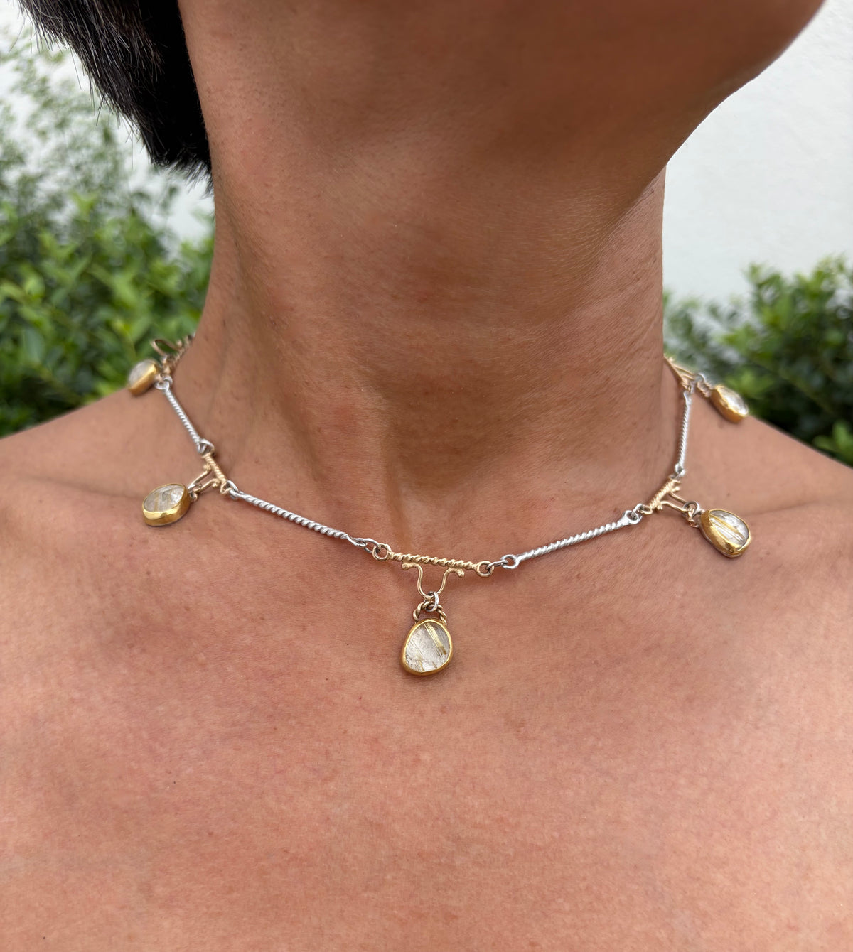 A close-up of a person's neck and upper chest wearing a delicate necklace with a silver and gold twisted link chain, and several small teardrop-shaped stones in gold bezels.