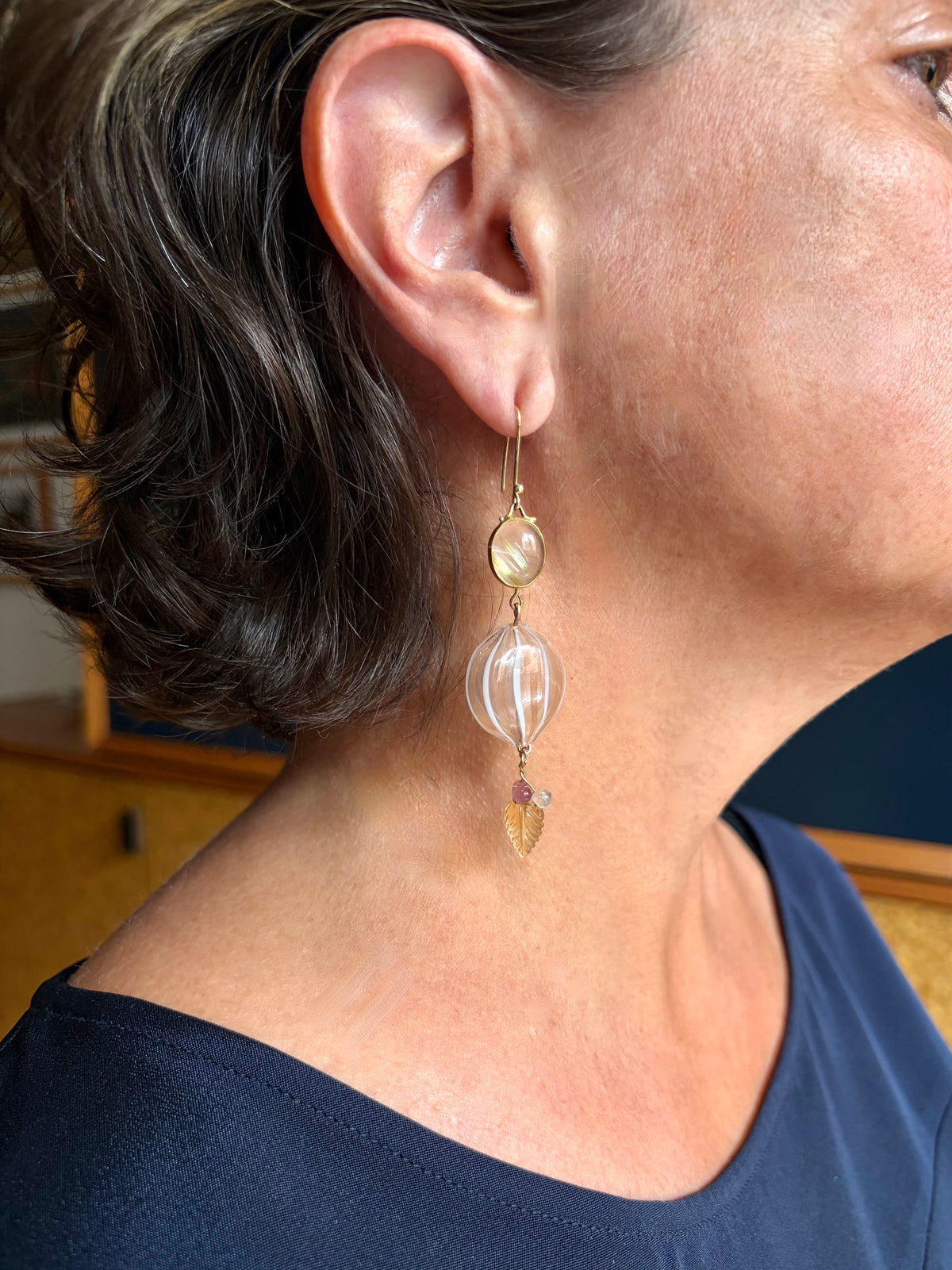A close-up of a person's ear and neck wearing a long dangling earring. The earring has a gold hook, a small oval quartz set in gold, and a larger round blown glass bead with metallic pink and white stripes. It is finished with a cluster of yellow, pink, and clear gemstones.