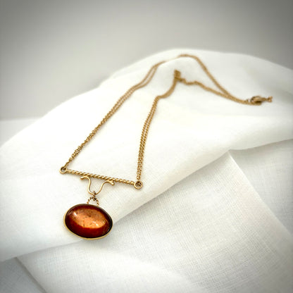 Gold necklace with an oval orange stone in a gold bezel hanging from a twisted gold pendant, on a delicate gold chain. The necklace lays over a white linen fabric.  