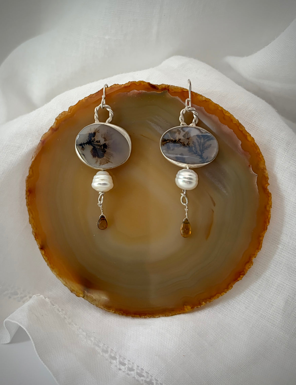 A pair of silver earrings featuring east-west ovals of dendritic agate that looks like seaweed floating in the sea. They are adorned with baroque South Sea pearls and delicate amber tourmaline drops. The pair rests on a cream colored slice of agate on white fabric.