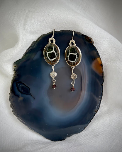 Silver earrings with oval brown, black, and white gemstones with quartz and garnet gemstones dangling beneath. They sit on a dark blue agate stone on a white fabric background. 