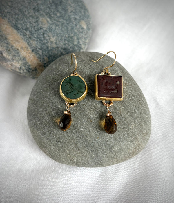 Pair of earrings with animals carved into gemstone surfaces, one in green and the other brown. A smokey quartz briolette dangles from each. They are positioned on a smooth beach stone, on a white linen ground. 