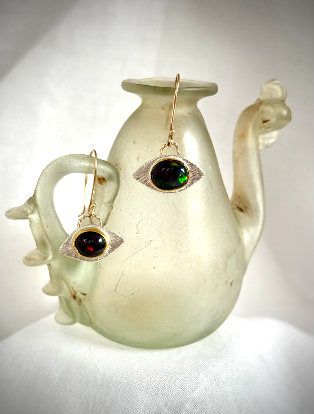 Pair of earrings in an eye design with a dark opal oval stone with flashes of green and red. They hang on the lip of  a white Roman glass vessel.