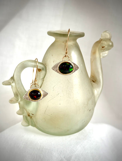Pair of earrings in an eye design with a dark opal oval stone with flashes of green and red. They hang on the lip of  a white Roman glass vessel.