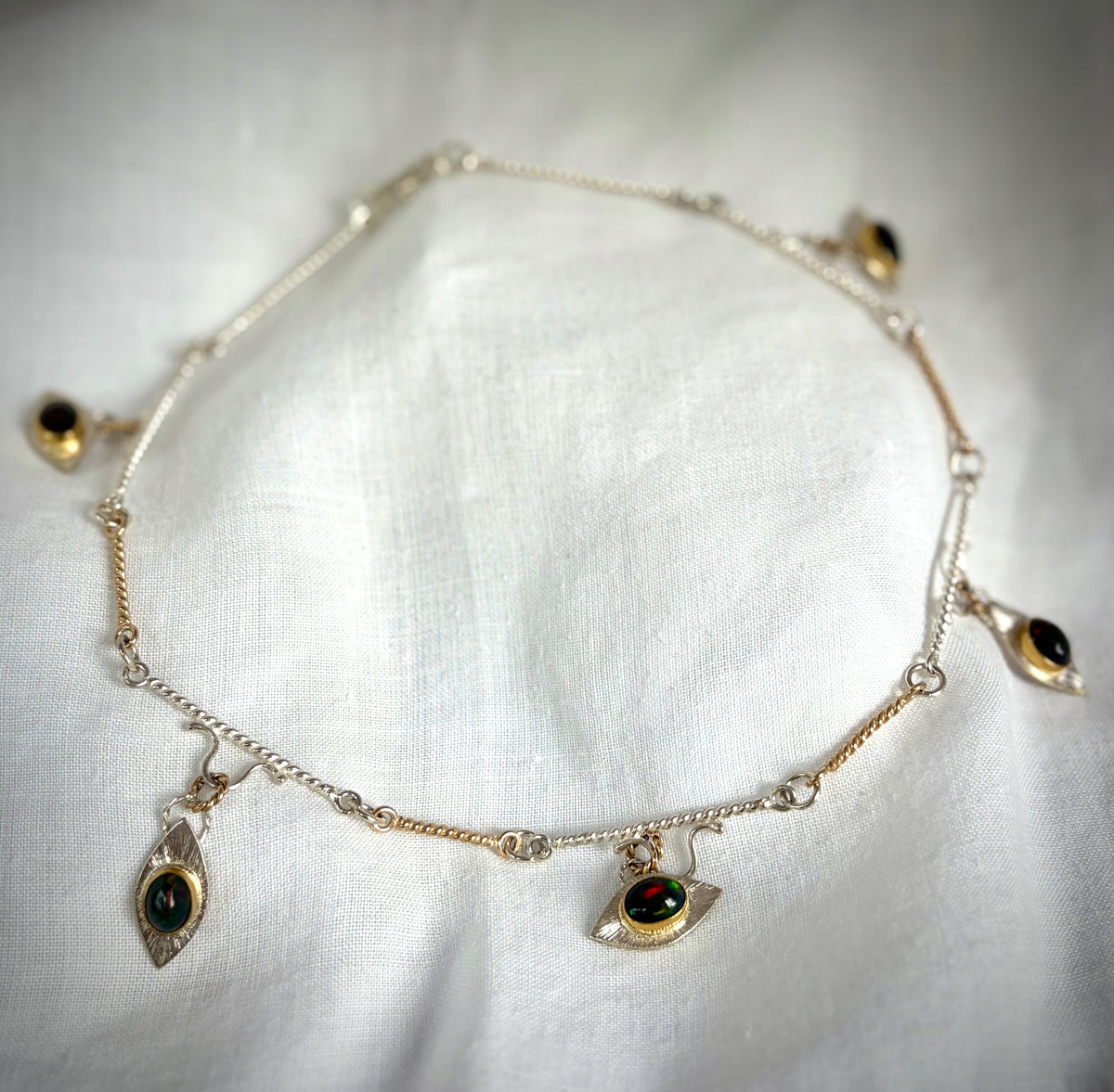A gold and silver link necklace with eye-shaped pendants on a white fabric background. At the center of each eye is a dark stone with green and gold flashes of color.