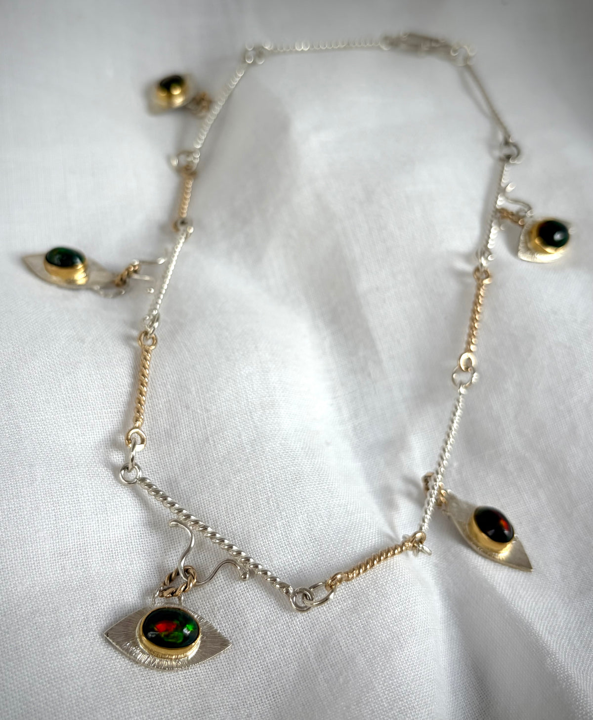 A gold and silver link necklace with eye-shaped pendants on a white fabric background. At the center of each eye is a dark stone with green and gold flashes of color.