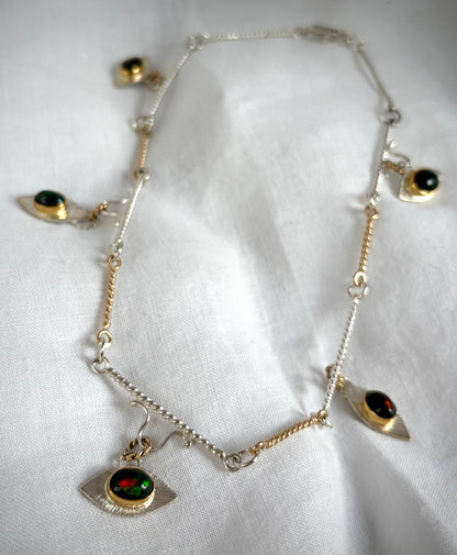 A gold and silver link necklace with eye-shaped pendants on a white fabric background. At the center of each eye is a dark stone with green and gold flashes of color.