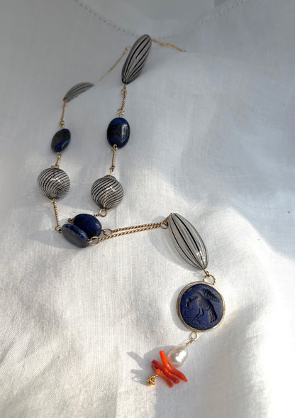 A necklace twists diagonally across a white fabric-covered surface. The twisted gold wire is punctuated by large, oval, clear glass beads with black stripes and round ones with swirling parallel lines. In between are gold-flecked lapiz lazuli beads, with a focal pendant that features a lapis carved with a bird with a snake in its beak. A South Sea baroque pearl, natural coral, and tiny gold beads complete the necklace.