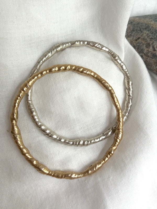 Two bangles, one gold, and the other silver on a white fabric with a sea stone in the background