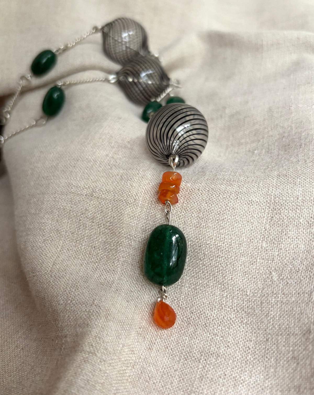 A close up view of a necklace with black-striped hand blown glass beads, deep green, smooth, emerald beads, and orange carnelian beads on silver hand-forged links. It sits on a flax-colored fabric background