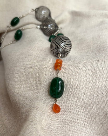 A close up view of a necklace with black-striped hand blown glass beads, deep green, smooth, emerald beads, and orange carnelian beads on silver hand-forged links. It sits on a flax-colored fabric background