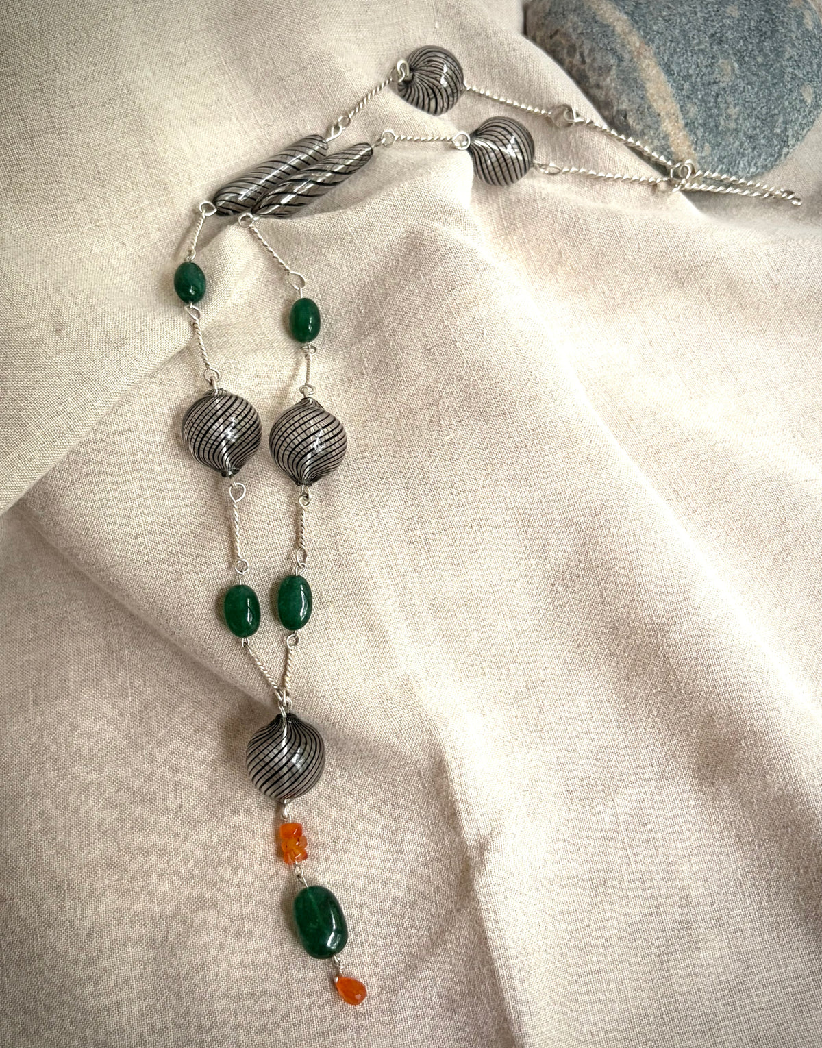 An overhead view of a necklace with black-striped hand blown glass beads, Zambian emerald beads, and orange carnelian beads on silver hand-forged links. It sits on  flax-colored fabric with a grey rock in the background