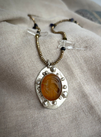 A beaded necklace with a pendant featuring a bird carved into a yellow-orange stone set in silver. The necklace rests on a textured fabric background