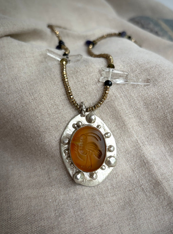 A beaded necklace with a pendant featuring a bird carved into a yellow-orange stone set in silver. The necklace rests on a textured fabric background
