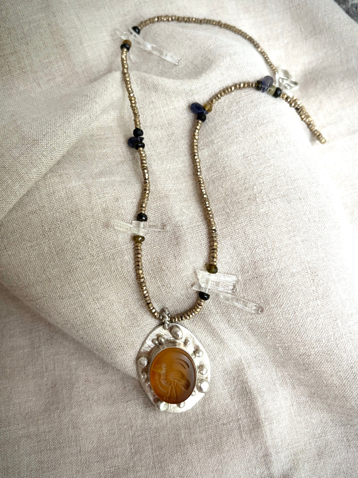 A beaded necklace with a pendant featuring a bird carved into a yellow-orange stone set in silver. The necklace rests on a textured fabric background