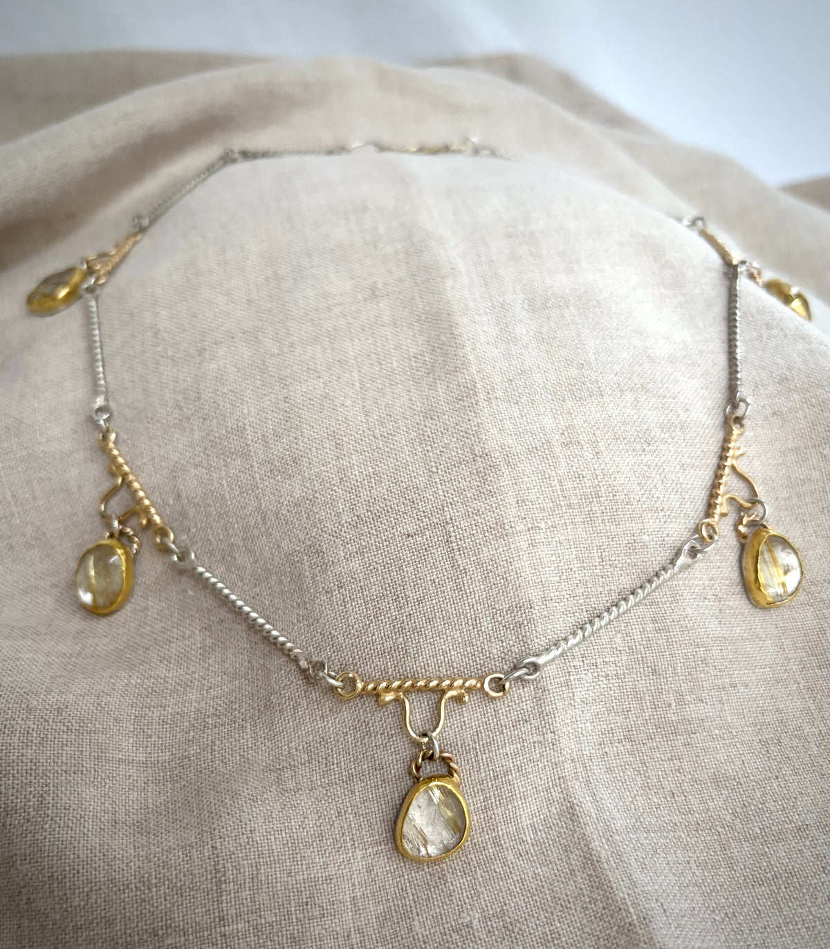 Gold and silver necklace with five golden rutile  quarts charms resting on a flax-colored fabric background