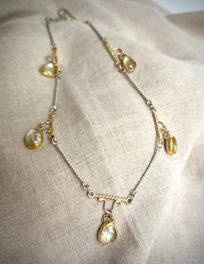 Gold and silver necklaces with five golden rutile  quartz charms resting on a flax-colored fabric background