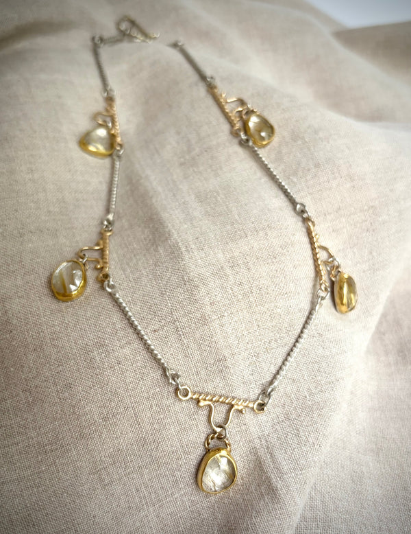 Gold and silver necklaces with five golden rutile  quartz charms resting on a flax-colored fabric background