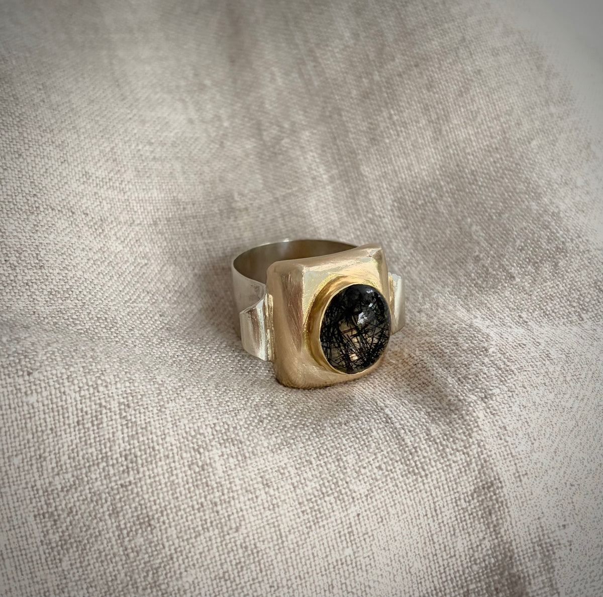 Silver scrolled band with a gold focal point set with a clear quartz gemstone with black rutile. It sits on a textured flax-colored background