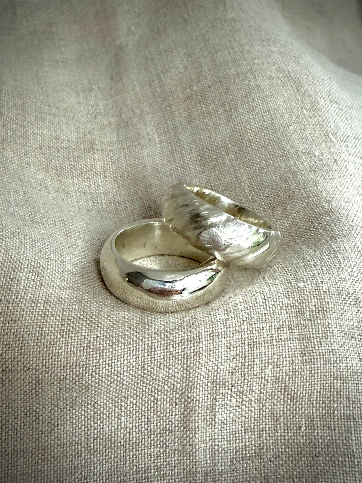 Two chunky silver bands, one smooth and shiny, and the other with a textured design on a flax-colored fabric background.