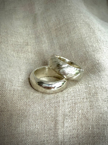 Two chunky silver bands, one smooth and shiny, and the other with a textured design on a flax-colored fabric background.