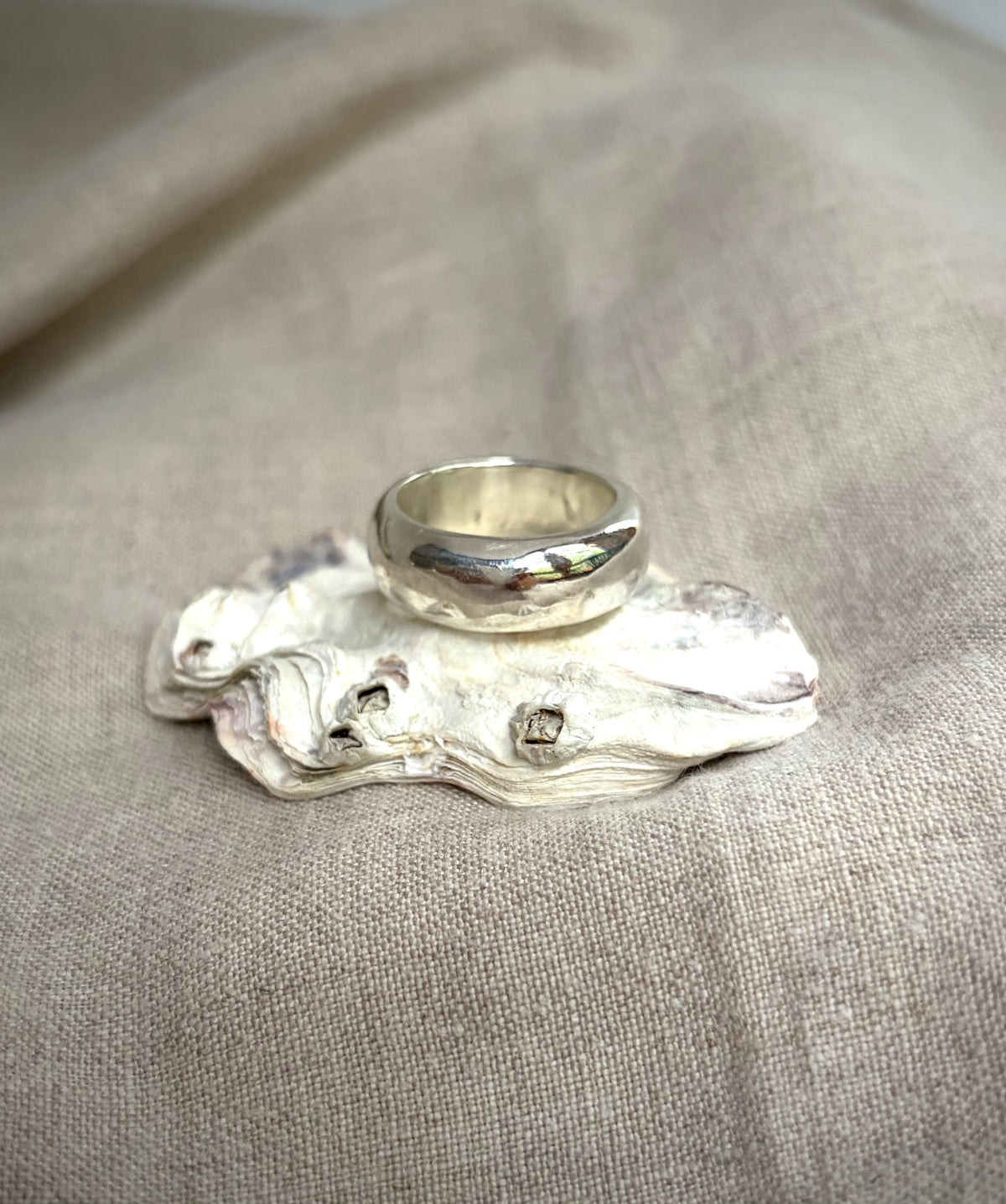 A chunky, shiny silver band sitting on a shell, on a textured beige surface