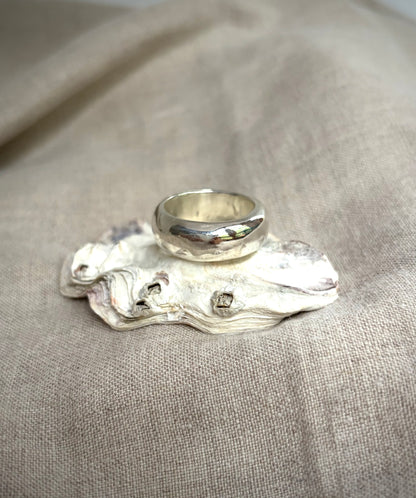 A chunky, shiny silver band sitting on a shell, on a textured beige surface