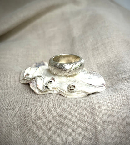 A chunky, textured silver band sitting on a shell, on a textured beige surface