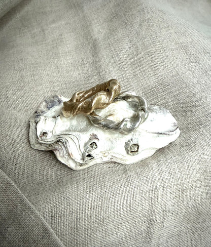 One gold twsited metal ring sits against another silver twisted ring, on top of a white oyster shell. There is a flax-colored fabric in the background. 