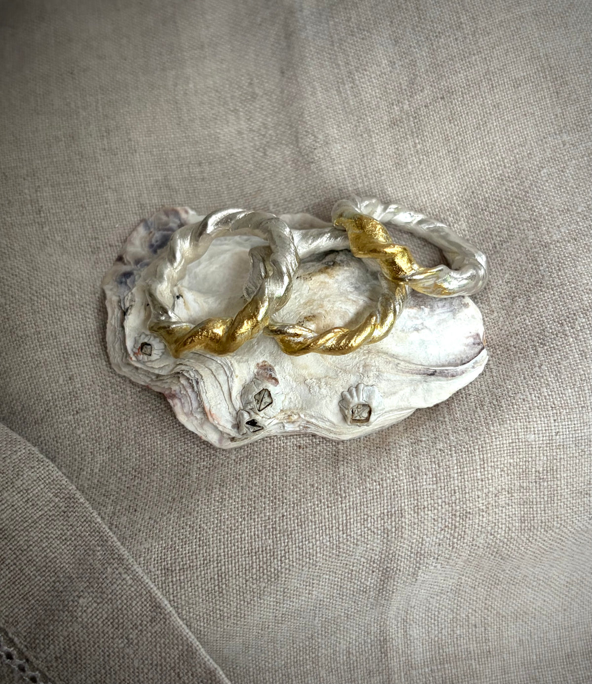 Three silver rings made of twisted metal sit on a blue agate stone slice. All three of the rings have a gold accent r. They sit on an oyster shell on a flax colored fabric background. 