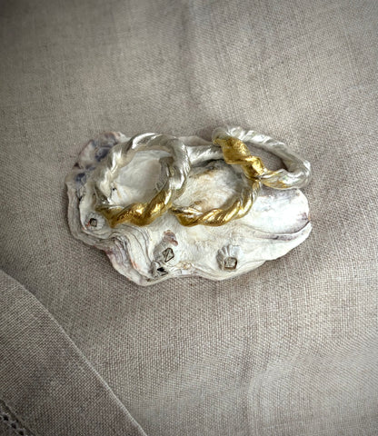 Three silver rings made of twisted metal sit on a blue agate stone slice. All three of the rings have a gold accent r. They sit on an oyster shell on a flax colored fabric background. 