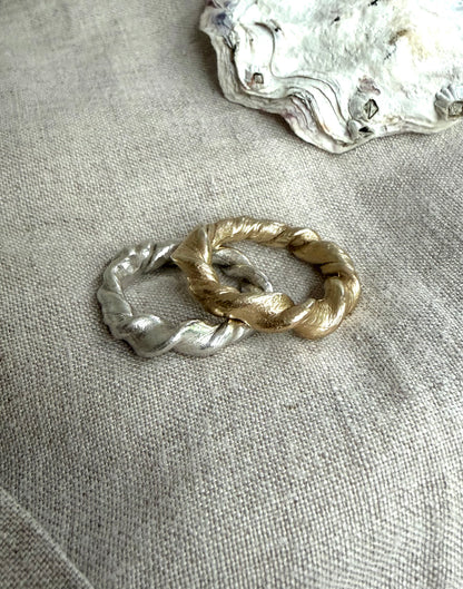 A pair of silver and gold twisted rings on a textured flax-colored fabric background. There is an oyster shell at the upper right.