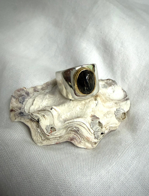 Silver ring with a clear quartz gemstone run through with black rutile set with a gold bezel. It sits on a white sea shell on top of a white textile. 