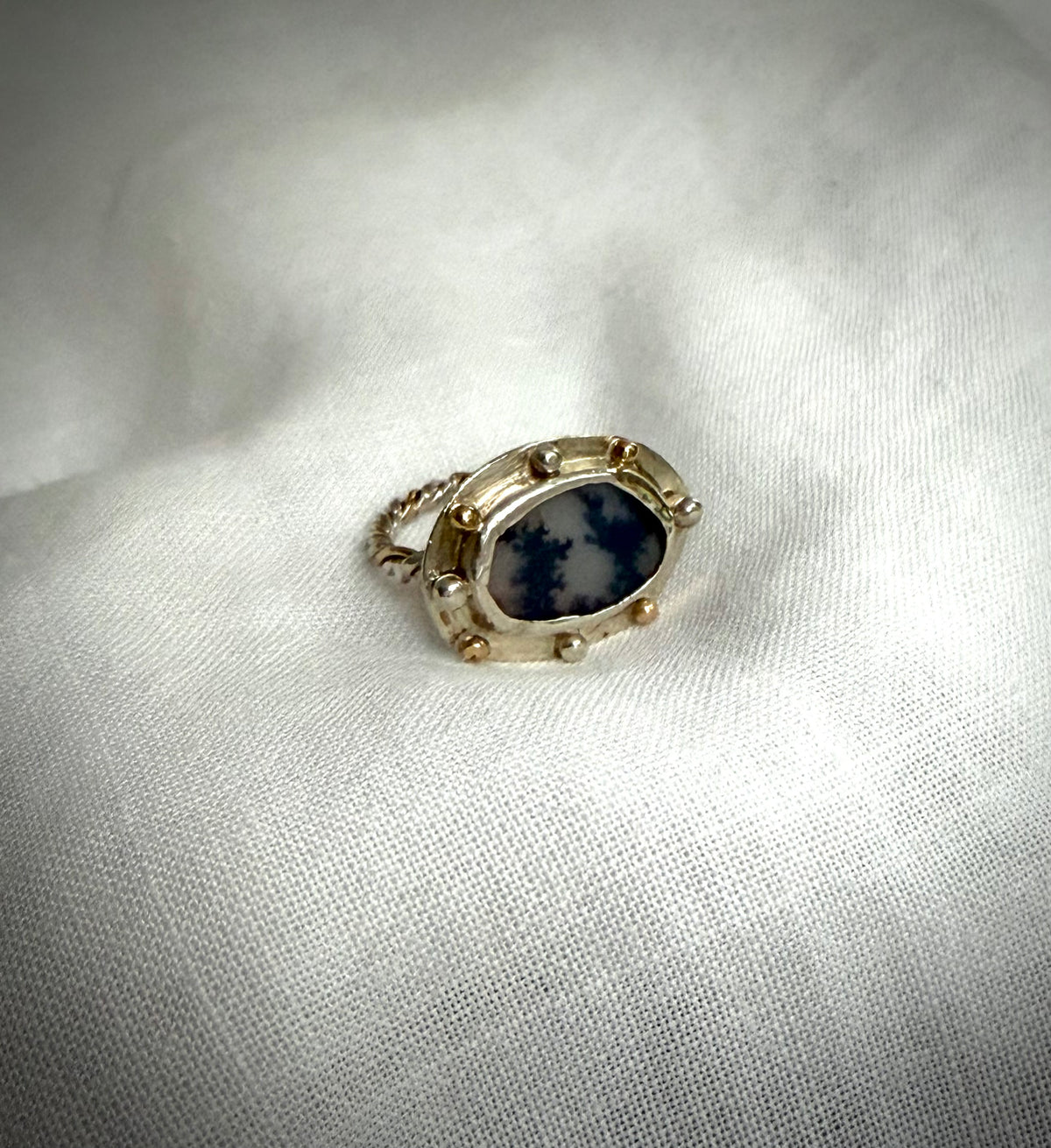 An oval ring-face featuring a dendritic agate which appears to have two trees silhouetted against a changeable grey sky. It is set in a silver surround with gold and silver granules and with a gold and silver twist band. It sits white piece of fabric.