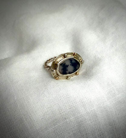 An oval ring-face featuring a dendritic agate which appears to have two trees silhouetted against a changeable grey sky. It is set in a silver surround with gold and silver granules and with a gold and silver twist band. It sits white piece of fabric.