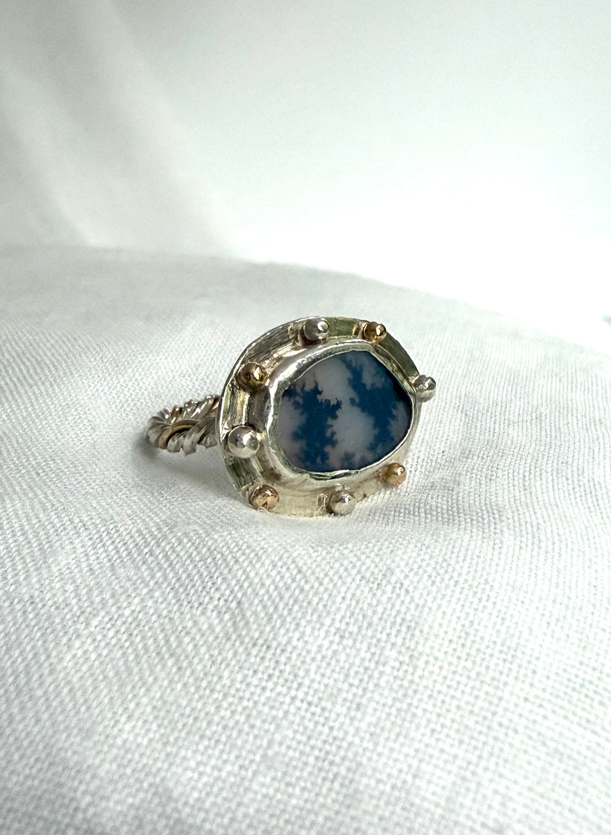 An oval ring-face featuring a dendritic agate which appears to have two trees silhouetted against a changeable grey sky. It is set in a silver surround with gold and silver granules and with a gold and silver twist band. It sits white piece of fabric.