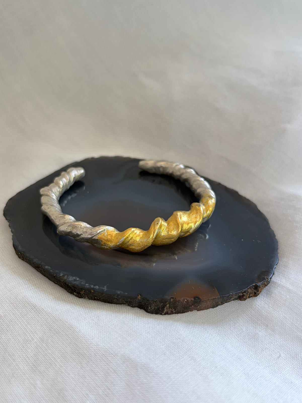A twisted silver cuff bracelet with alternating textured gold and silver sections, displayed on a dark polished stone slice against a light fabric backdrop.