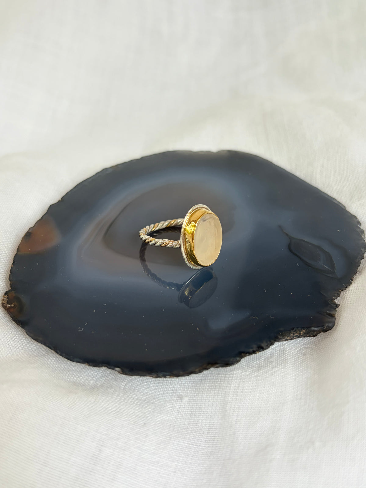 A gold and silver ring with an oval bezel-set stone displayed on a polished navy blue agate slice, set against a white fabric background. The band of the ring is twisted gold and silver wire. 