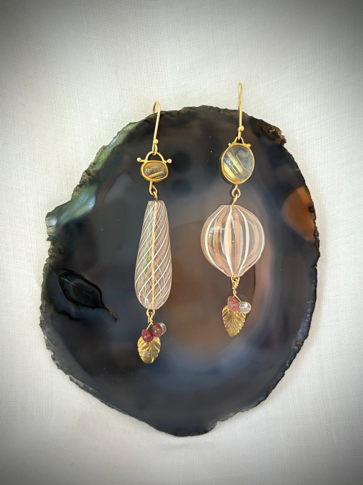 A pair of gold-tone dangle earrings displayed on a polished dark agate slice. The earrings feature striped glass beads in pale pink and cream, small oval clear stones with gold stripes, and gold leaf-shaped stones with tiny pink gem accents.