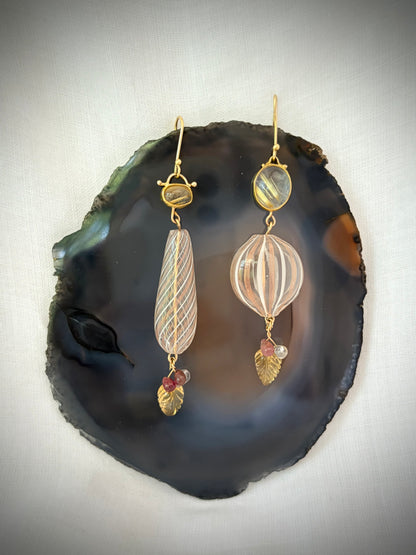 A pair of gold-tone dangle earrings displayed on a polished dark agate slice. The earrings feature striped glass beads in pale pink and cream, small oval clear stones with gold stripes, and gold leaf-shaped stones with tiny pink gem accents.