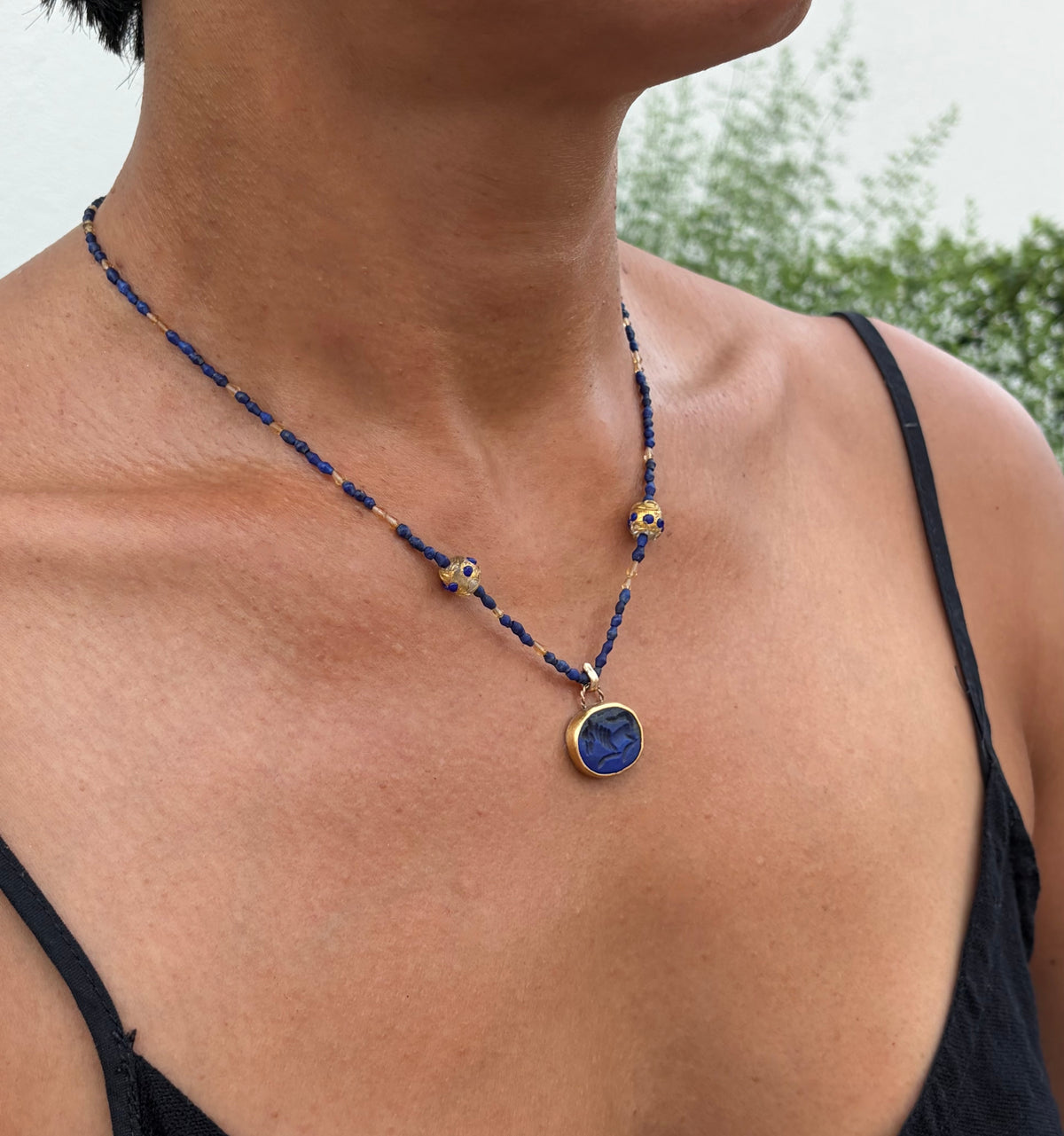 A close-up of a person wearing a beaded necklace with a round blue pendant featuring a horse design, set against a blurred outdoor background.
