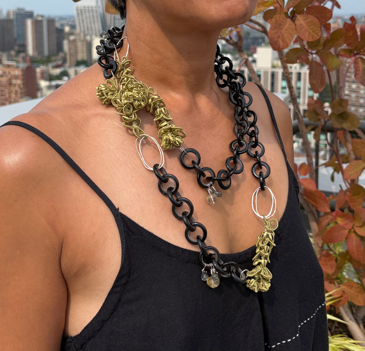 A close-up lifestyle shot of a model wearing a multi-strand statement necklace. The necklace features black chain links, textured chartreuse silk jasmine-like charms, silver and gold links, and small quartz beads and is doubled around the model’s neck. 