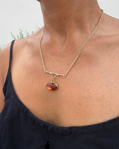 A gold chain necklace with a twisted gold pendant dangles a cabochon pendant in warm amber, red, and orange tones, shown worn on a model's neck. 