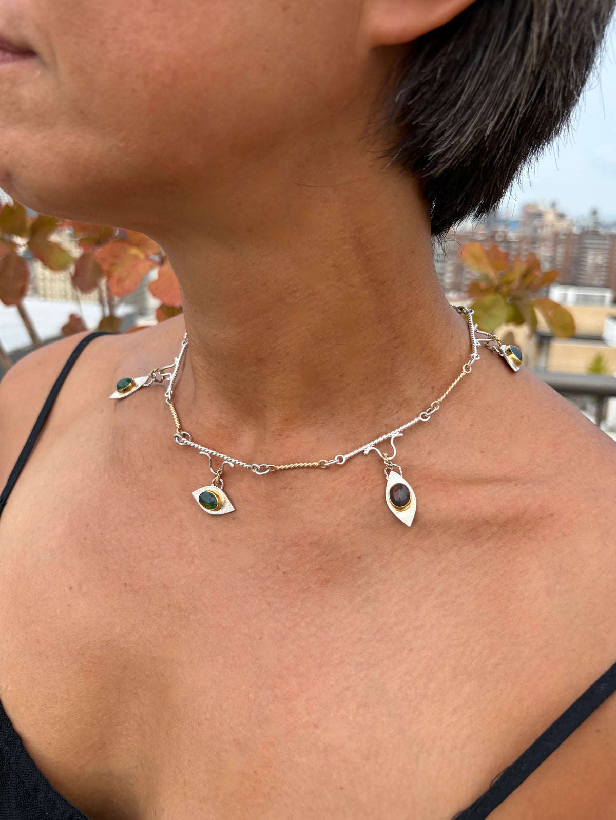 A close-up lifestyle shot of a person wearing a silver and gold chain necklace with dangling eye-shaped charms, each containing a dark oval stone with green, orange, and red flashes of color.