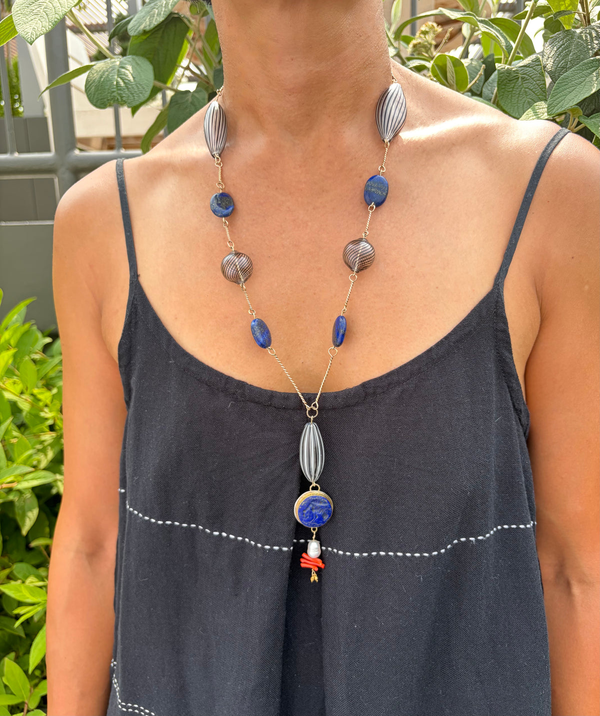 A person wearing a long beaded necklace with lapis beads and black-striped glass beads, styled with a black camisole top against a background of green foliage.