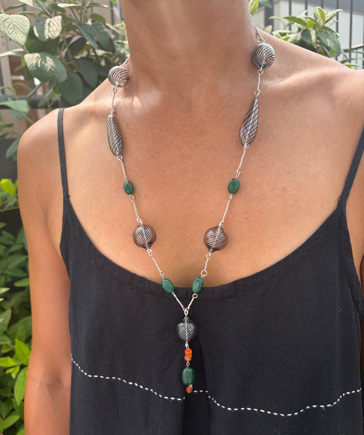 A necklace with black-striped hand-blown glass beads, green emerald beads, and orange carnelian bead accents on silver hand-forged links. It is worn by a model in a black sundress against a background of green foliage.