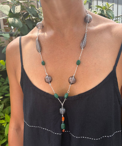 A necklace with black-striped hand-blown glass beads, green emerald beads, and orange carnelian bead accents on silver hand-forged links. It is worn by a model in a black sundress against a background of green foliage.