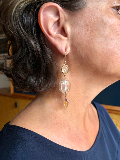 A close-up of a person's ear and neck wearing a long dangling earring. The earring has a gold hook, a small oval quartz set in gold,  and a larger round blown glass bead with metallic pink and white stripes. It is finished with a cluster of yellow, pink, and clear gemstones. 