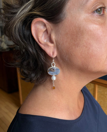 A close-up of a woman's ear wearing a dangling earring. The earring features a silver hook, an oval stone with natural inclusions, a white pearl, and a small amber-colored tourmaline gemstone.